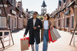 interracial couple in trendy coats and sunglasses walking with shopping bags and looking away on bridge.