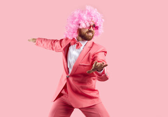 Funny bearded man in wig, glasses and suit having fun isolated on pink background. Caucasian man in pink wig and coral formal suit is having fun dancing. Holiday fun and carnival concept.