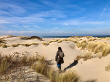 Rear View Of Woman And Her Shadow Walking In A Dunescape, The Sea At The Horizon, The Sky Is Blue