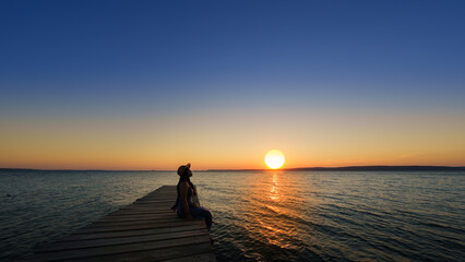 Wall Mural - Traveler asian woman with her summer vacations on a twilight by the sea her sitting on bridge lonely dramatic lifestyle. Aegean Sea Beach, Canakkale, Turkye. Leave a blank and space for text entry.