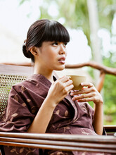 A Young Asian Woman In A Robe Drinking Tea On The Balcony Of An Open-air Spa Villa. Ubud, Bali, Indonesia.