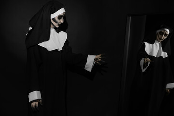 Wall Mural - Scary devilish nun near mirror on black background. Halloween party look