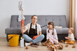 © sementsova321 - Young adult female sitting on floor among mess with her children, woman holding mop, doing domestic chores, cleaning apartment, looks tired, feels fatigue.
