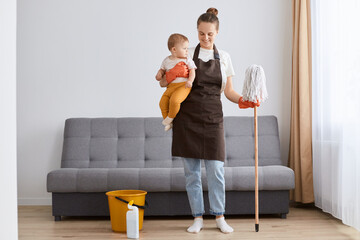 Wall Mural - Full length portrait of smiling joyful woman in apron washing floor with mop at home, doing domestic chores, cleaning apartment, holding infant baby in hands.