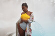 © TrueFrame Collective - Medium long portrait of beautiful African American woman wearing swimsuit, cover-up and sunglasses standing against thermal spring pool