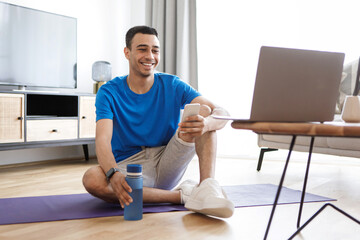 Online workout. Positive arab man exercising at home, looking at laptop, using smartphone, having fitness class online