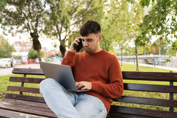 A young businessman outdoors in park sitting with his laptop talking on phone enjoying outside 