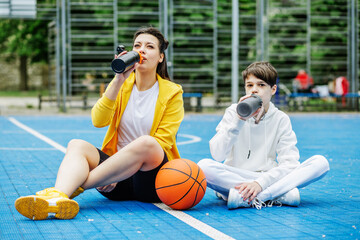 Wall Mural - Teenager boy and girl are sitting on sports court, drinking water and resting during break.