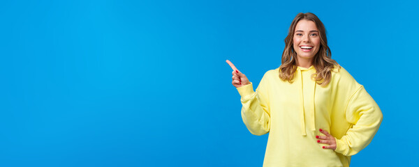 Take a look. Upbeat good-looking blond girl pointing finger left, recommend promo, advertise product, look pleased and smiling camera, explain project, standing blue background