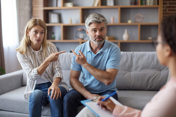 Wall Mural - Sad mature caucasian lady and male point fingers at each other in clinic office interior at meeting