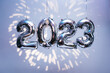 Silber foil balloons in numbers shape 2023 hanging against blue wall with fireworks projection. New Year holidays