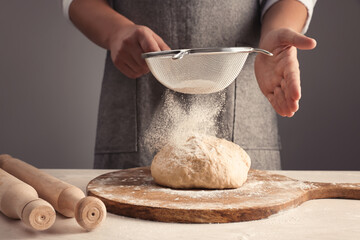 Man sprinkling flour over dough at table near grey wall, closeup