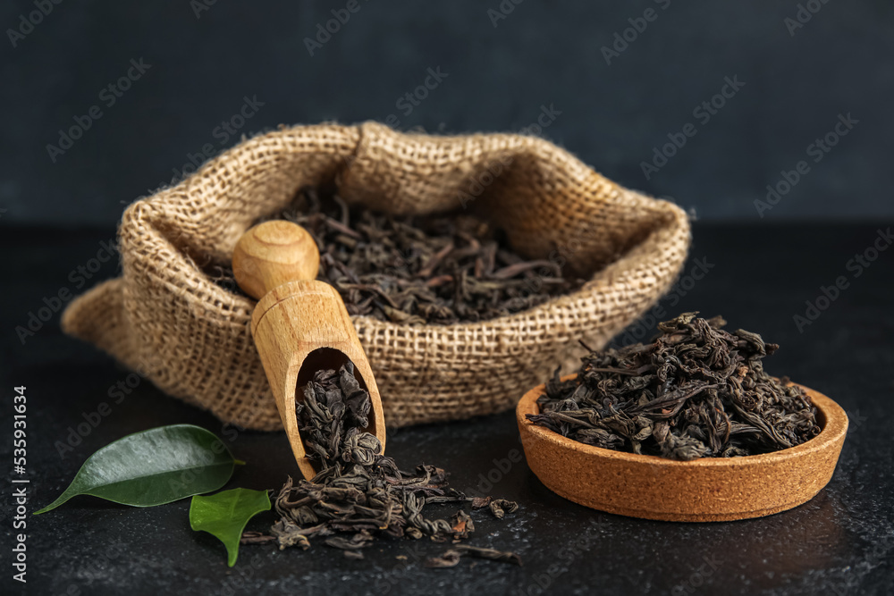Bag, bowl and scoop with dry tea leaves on dark background