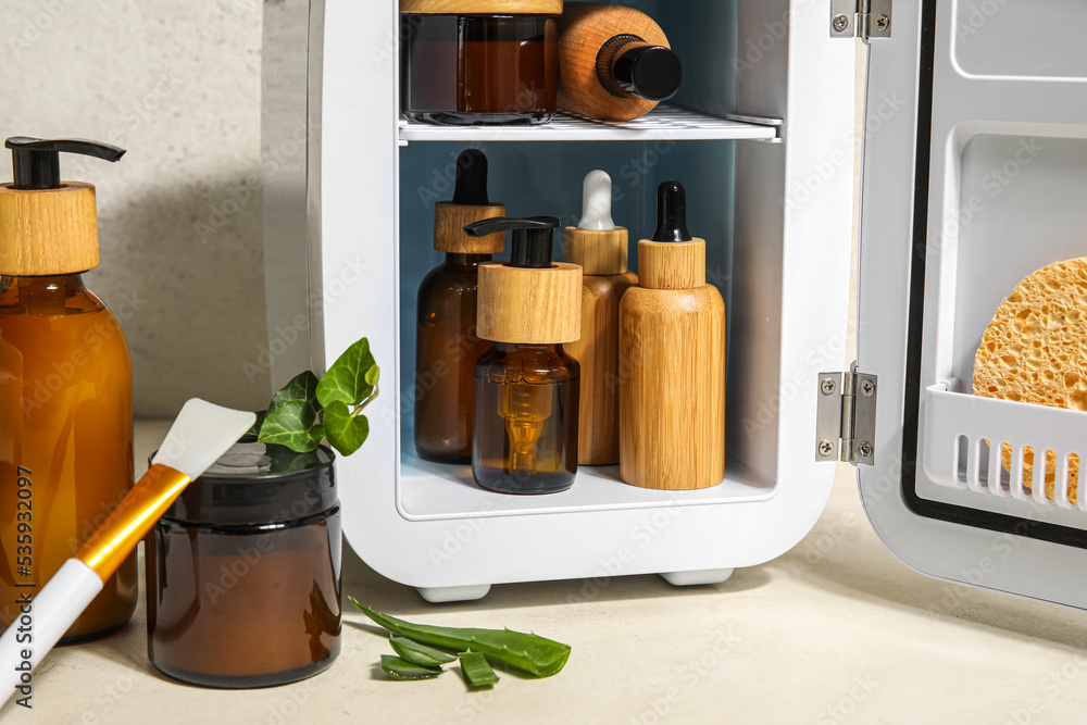 Small refrigerator with natural cosmetics, brush and plant leaves on light background