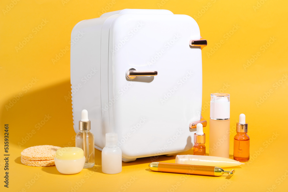 Small refrigerator with cosmetics and facial massage tool on yellow background