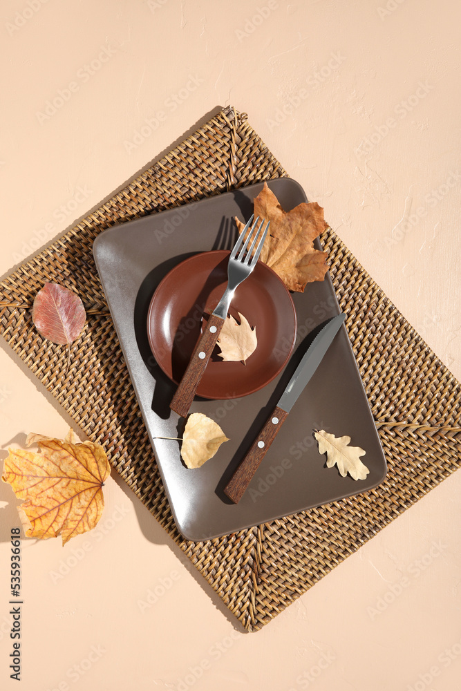 Autumn table setting with fallen leaves and wicker mat on beige background