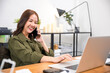 © sorapop - Happy Asian young woman working on laptop computer at home office while talking on mobile phone, smiling businesswoman having business call to talking with company sales client financial