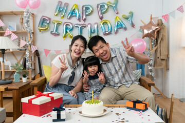 Wall Mural - happy asian small family looking at camera with victory hand gestures at the baby girl's second birthday celebration party at home with pink balloon and garland