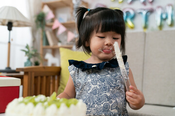 Wall Mural - closeup of happy asian toddler baby girl licking yummy whipped cream on face and looking playing with the cake knife on her second birthday party at home