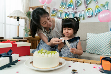 Wall Mural - smiling asian mother wiping baby girl's face with a tissue as she is playing with a cake server at the birthday party at home