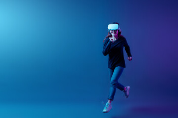 Asian woman wearing VR headset with experience playing video game and running and jumping levitating in the air on futuristic purple cyberpunk neon light background. Metaverse technology concept.