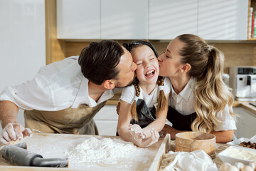 Adorable family in modern kitchen. father and mother kissing little daughter wearing aprons. Parents teaching daughter cooking baking kneading dough sieving flour preparing homemade pizza.