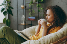Sideways Young Woman Of African American Ethnicity Wear T-shirt Drink Coffee Read Book Close Eyes Sits In Armchair Stay At Home Flat Rest Relax Spend Free Spare Time In Living Room Indoor Grey Wall.