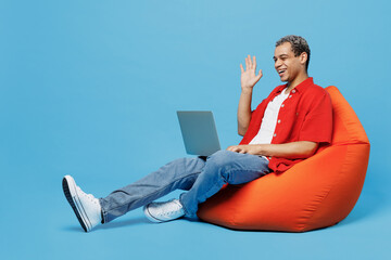 Wall Mural - Full body young IT man of African American ethnicity wear red shirt sit in bag chair hold use work on laptop pc computer waving hand get video call isolated on plain pastel light blue cyan background.