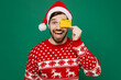 © ViDi Studio - Merry young man 20s in red warm knitted sweater Santa hat posing hold in hand cover eye with credit bank card isolated on plain dark green background studio New Year 2023 holiday celebration concept