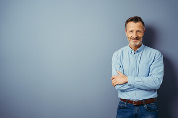 Wall Mural - Handsome middle-aged man, standing with his arms folded, looking at camera and smiling. Half-length front portrait against blue background with copy space