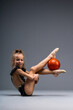 A young girl gymnast in a gymnastic leotard does exercises with a ball. Isolated on a gray background.