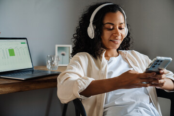 Wall Mural - Black young woman in headphones using mobile phone at home