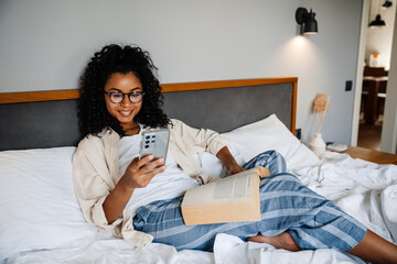 Wall Mural - Black young woman using mobile and reading book on bed at home