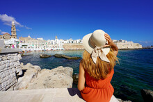Young Traveler Woman In Orange Dress And Hat Sitting On The Quay Of Monopoli Port Looking The Historic Town From Seaview. Travel Destination In Apulia Region, Italy.