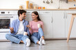 © Prostock-studio - Cheerful smiling young wife and husband with cups of hot drink sitting on floor and talking at kitchen