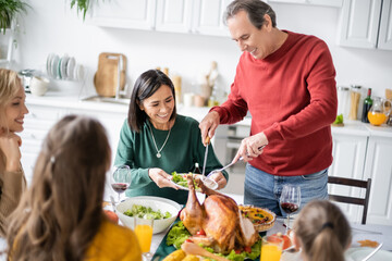 Wall Mural - Cheerful grandparent putting turkey on plate near multicultural women and kids during thanksgiving dinner