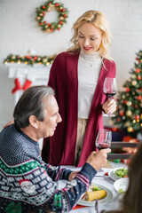 Wall Mural - happy blonde woman with glass of red wine toasting near senior father during christmas dinner in living room