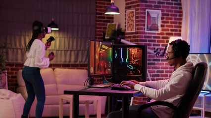 Wall Mural - Gamer playing multiplayer first person shooter while gaming girl is fighting in virtual reality boxing game. Man streaming online action game on pc while girlfriend uses vr goggles for simulation.