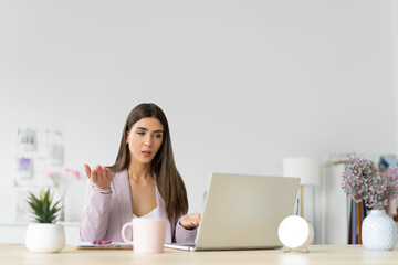 Outraged and surprised young woman in a pink suit in front of a laptop screen. Bright home office. Copy space.