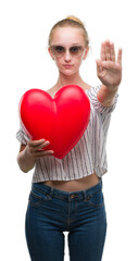 Poster - Blonde teenager woman holding red heart with open hand doing stop sign with serious and confident expression, defense gesture