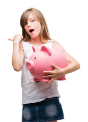 Sticker - Young blonde toddler holding piggy bank pointing with hand and finger up with happy face smiling