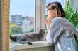© Valerii Honcharuk - Mature smiling woman looking out window at home with relaxed cat