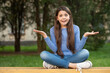 Outraged teenage girl shrugging shoulders sitting legs crossed on bench outdoors, outrage
