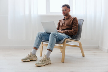 Smiling cheerful happy young man guy in eyewear looks at screen laptop enjoys chatting with friends doing cool NFT project sitting on chair at home. Distance communication Remote Work New profession