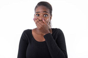 Wall Mural - Portrait of shocked African American woman covering mouth. Scared young model in black shirt with ponytail looking at camera with hand on mouth. Studio shot, shock concept.