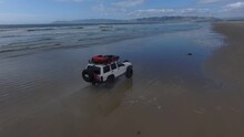 A White Jeep Cherokee Driving North Up The Beach And  Through The Waves Of The Pacific Ocean.  12 Seconds