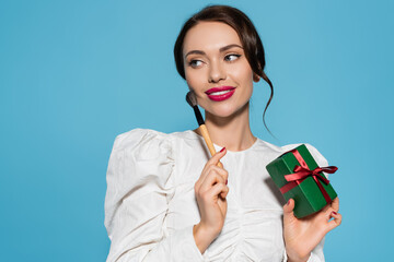 Canvas Print - cheerful young woman in white blouse holding wrapped present and cosmetic brush isolated on blue.