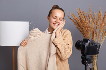 Wall Mural - Portrait of satisfied delighted young adult woman blogger stylist with bun hairstyle shooting video on camera on tripod, showing subscribers a warm soft sweater with a new collection.