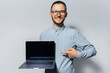 © Lalandrew - Studio portrait of young smiling man, holding laptop in hand, pointing index finger on screen, on white background. Wearing blue shirt and eyeglasses.
