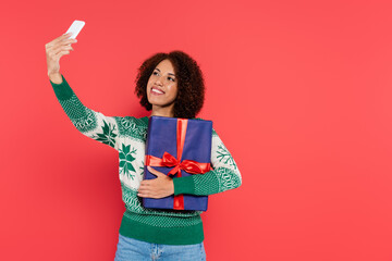 Wall Mural - happy african american woman in warm sweater with winter pattern taking selfie with blue gift box isolated on red